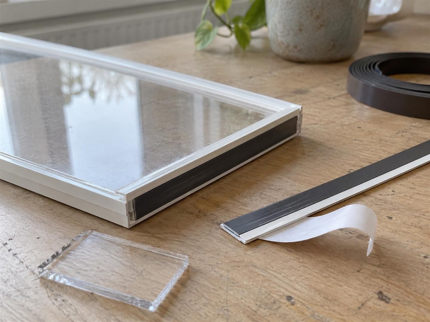 Hoek van een voorzetraam van helder kunststof in een smal wit profiel, met een magneetstrip en een losse stalen strip